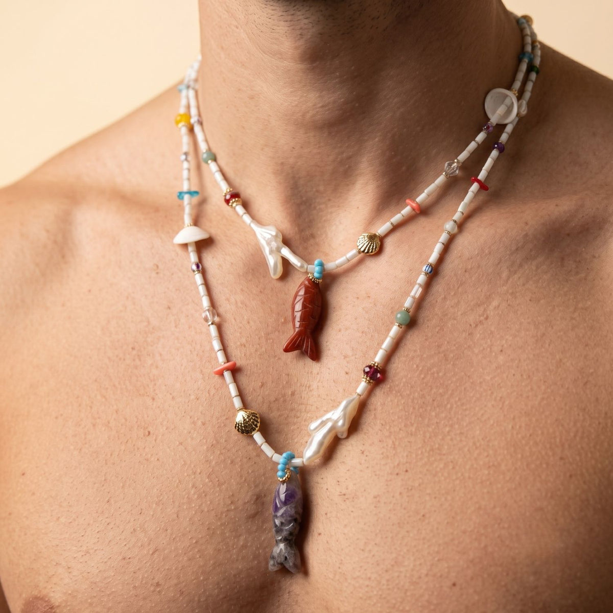 Close-up portrait of a male model wearing two layered Marisol Necklaces on a bare chest, showcasing the Red Jasper and Amethyst carved fish pendants, natural shells, and coral-shaped freshwater pearls adjusted to different lengths.