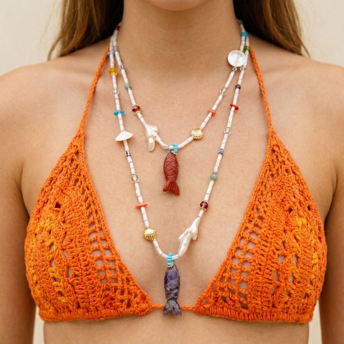 Front portrait of a female model wearing an orange crochet bikini top, featuring the Marisol Adjustable Necklace layered in both the Amethyst and Red Jasper fish pendant variations to show the sliding clasp adjustability, coral branch pearls, and natural shells.