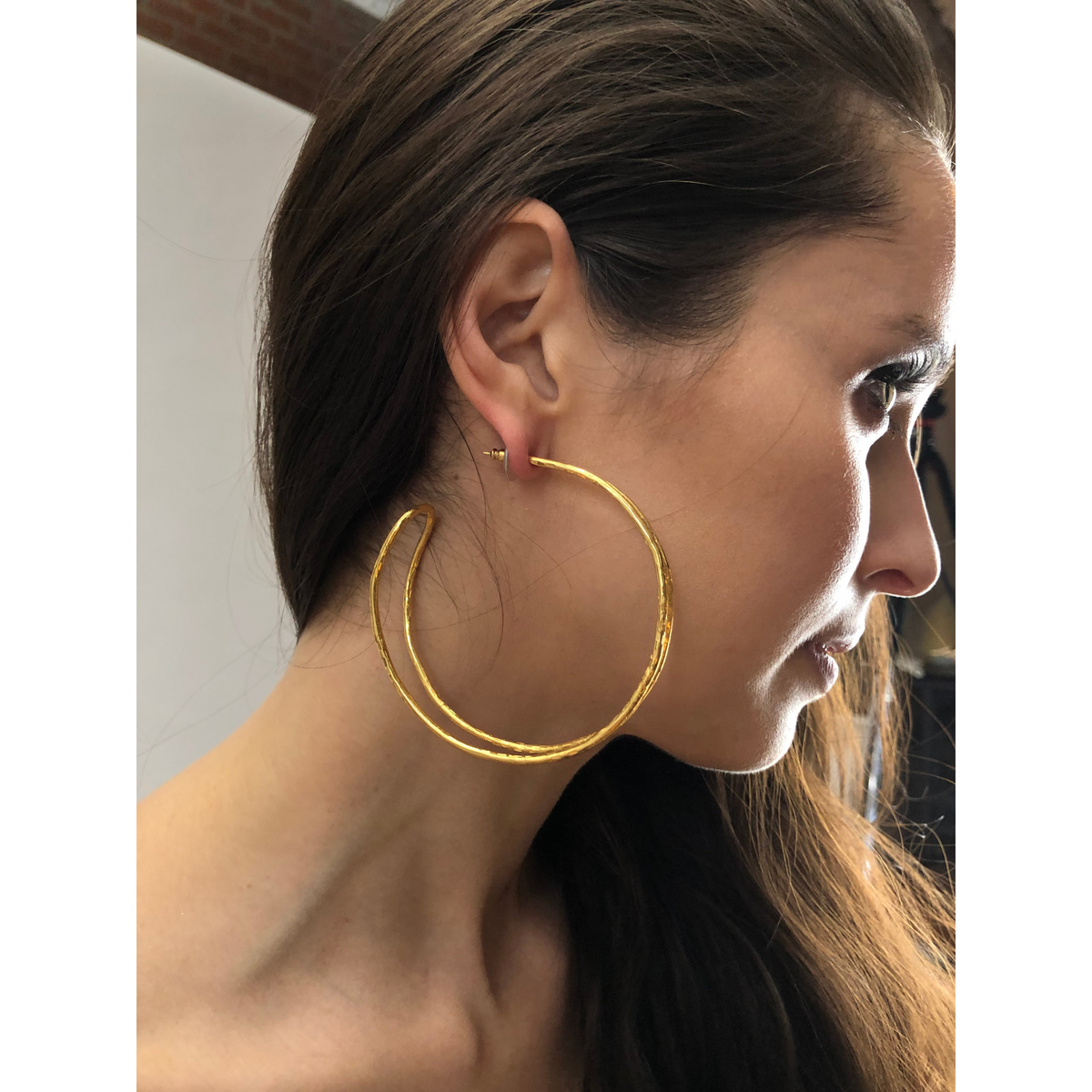 Un woman avec long brown hair wears JAREDJAMIN Bijoux en Ligne's or Mimi créole Post boucles d'oreilles, avec a large double-wire arch in 18K or plate et an incomplete circle design. She is shown in profile, facing right in natural light.