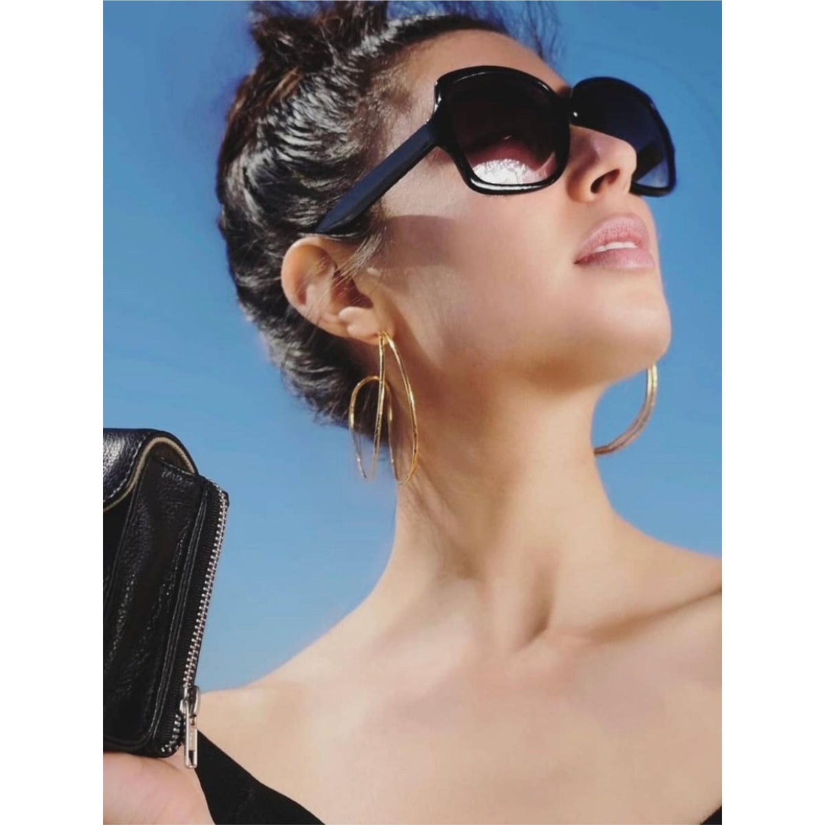 Un woman avec dark hair dans un bun wears large black sunglasses, JAREDJAMIN Bijoux en Ligne or Mimi créole Post boucles d'oreilles, et a black off-shoulder top, holding a black zippered clutch. Sunlight highlights her face as she looks up at the clear blue sky.