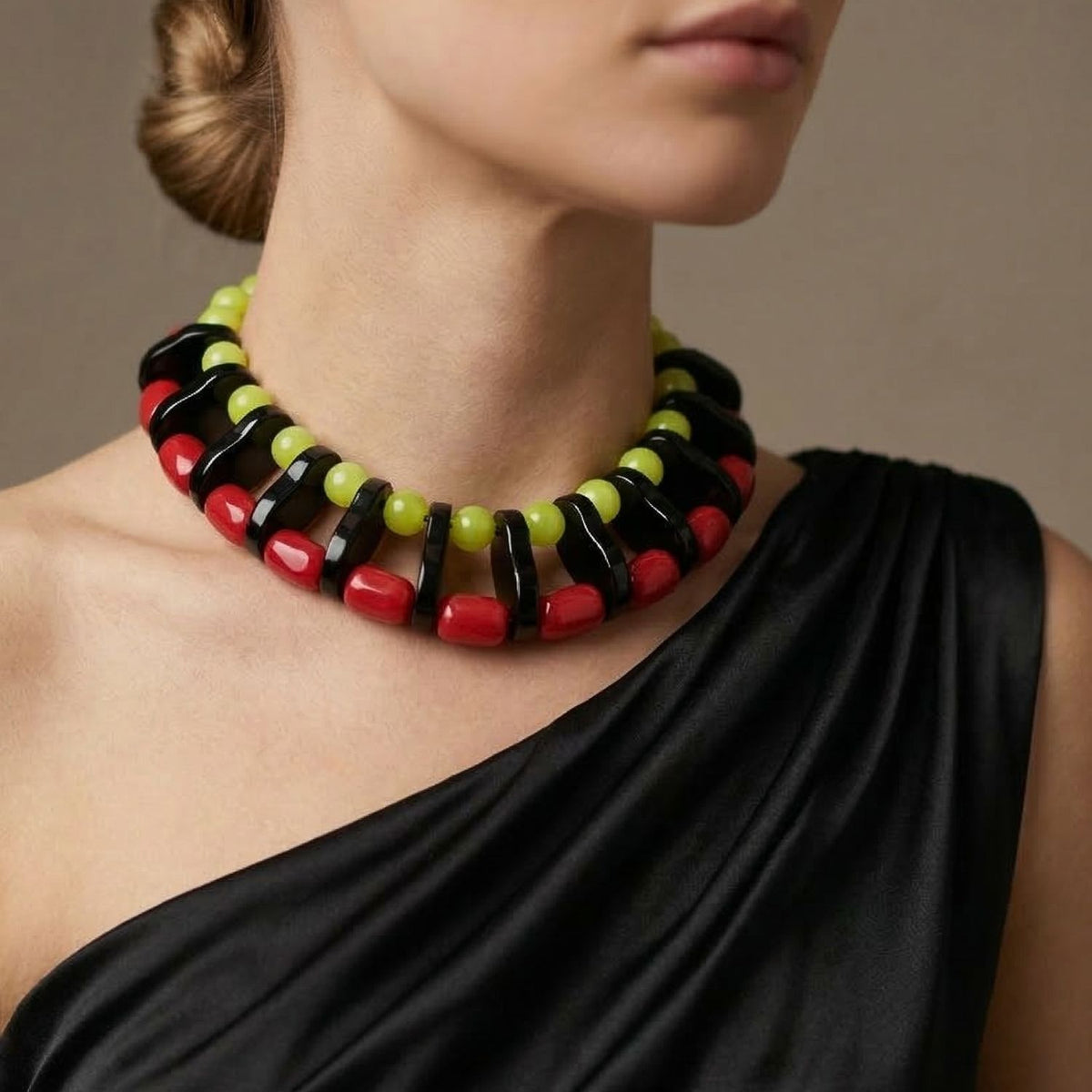 The Modern Mix Gemstone Necklace by JAREDJAMIN Jewelry Online, with striking red faceted beads and an adjustable extension, stands out against a woman’s black one-shoulder dress. The bold jewelry is highlighted by a neutral background.