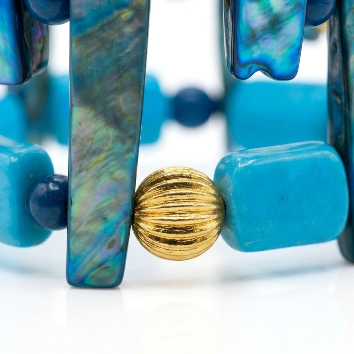 Macro studio detail shot focusing on the iridescence of the vertical dyed shell stick, smooth rectangular chalk turquoise bead, and ribbed gold-plated brass sphere.