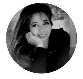 A grayscale portrait of a smiling woman with long, dark hair partially covering one side of her face. She rests her face on her hand, wearing a dark outfit. The background is softly blurred, and the image is circular in shape.