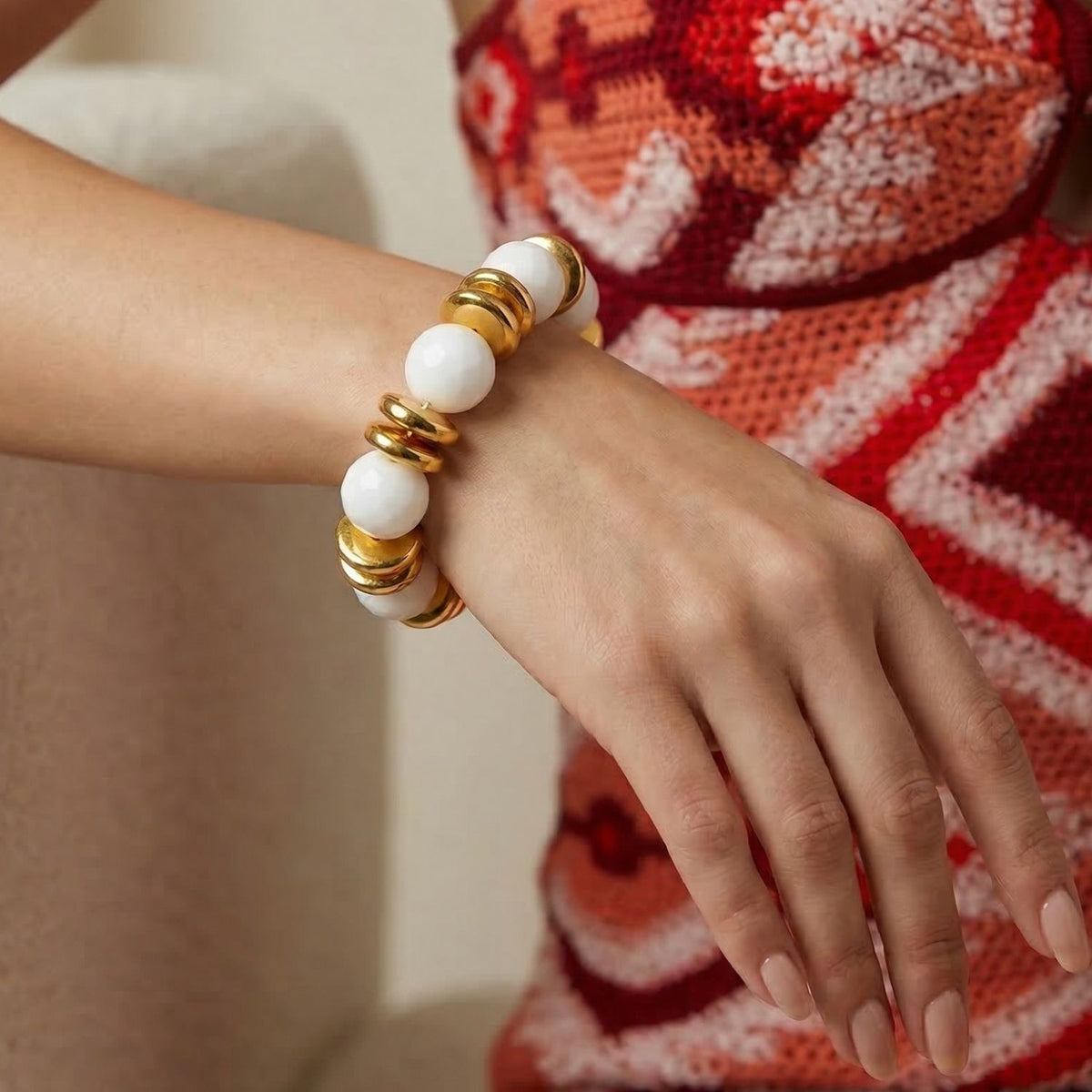 Los Olivia White Stretch pulsera de JAREDJAMIN Joyería Online—a chunky white agate pulsera con bañado en oro beads—is showcased sobre un person wearing a red, orange, y white crochet dress, their arm resting sobre un beige surface.