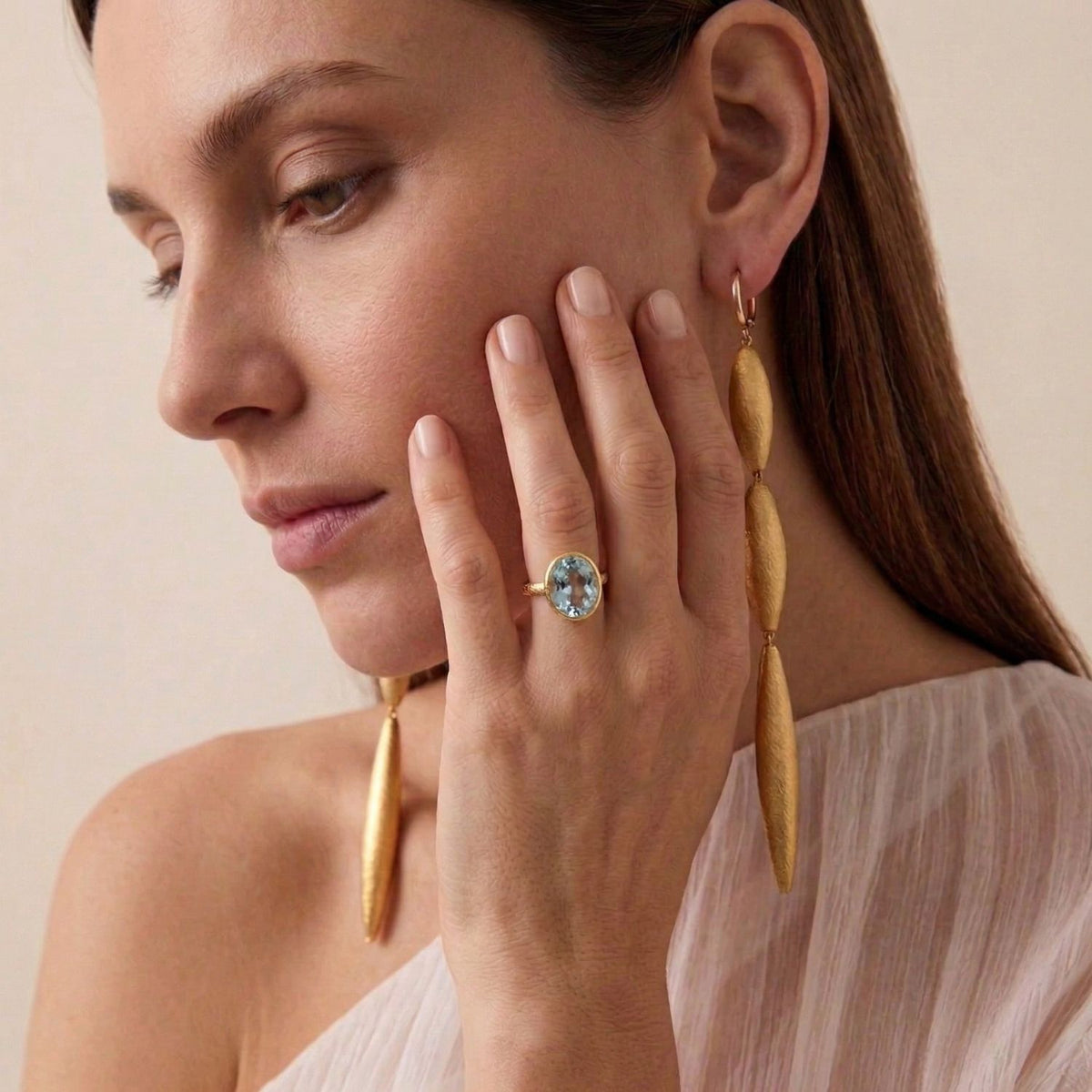 A woman with light skin and brown hair gazes down thoughtfully, wearing long gold leaf earrings and the JAREDJAMIN Jewelry Online Oval Aquamarine Gemstone Ring. Her hand rests near her face beneath a sheer, off-white one-shoulder top.