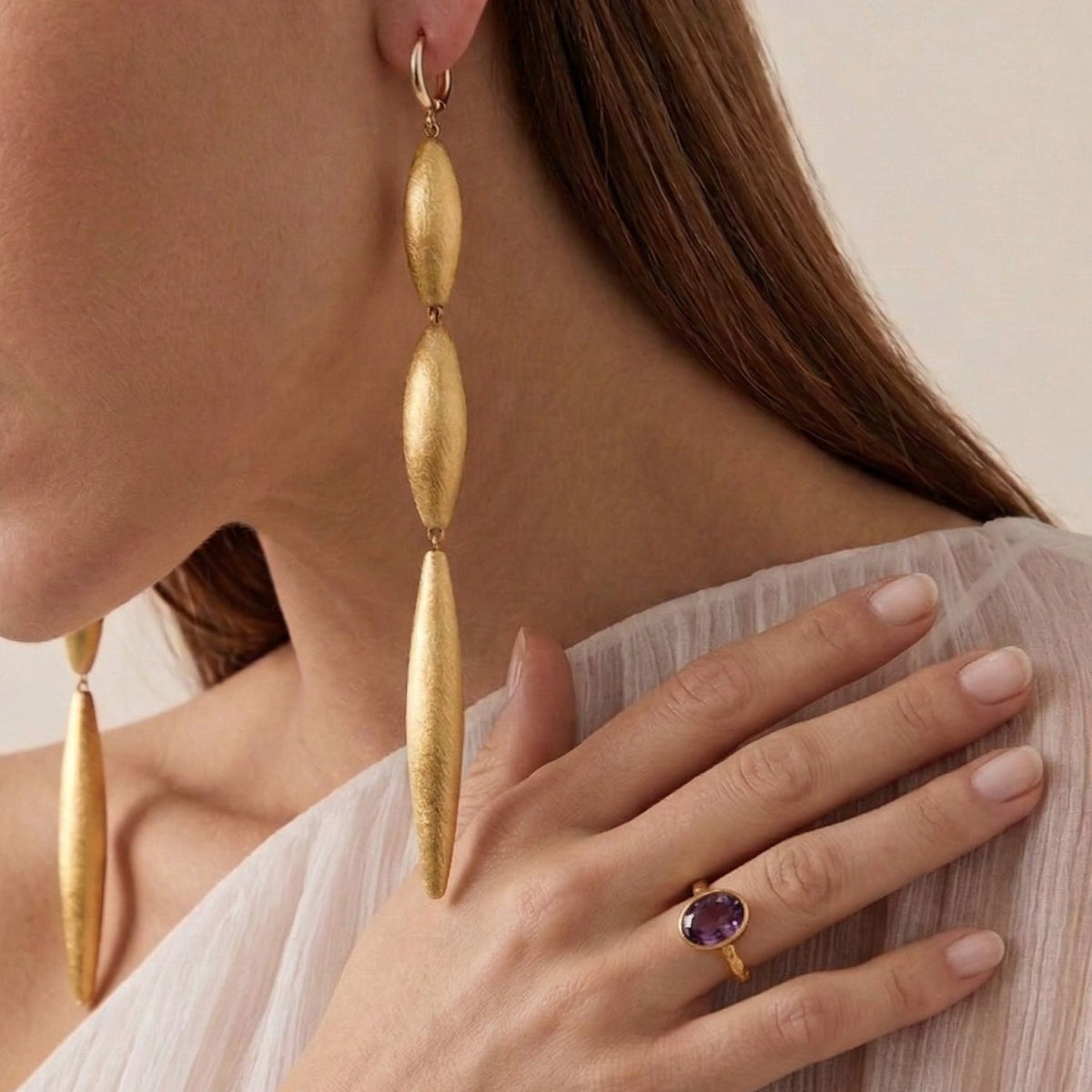 A woman wears long gold oval earrings and the JAREDJAMIN Jewelry Online Oval Amethyst Gemstone Ring, featuring an 18K gold-plated band and oval amethyst. She touches her shoulder, visible through sheer white fabric; only her hand and lower face are seen.
