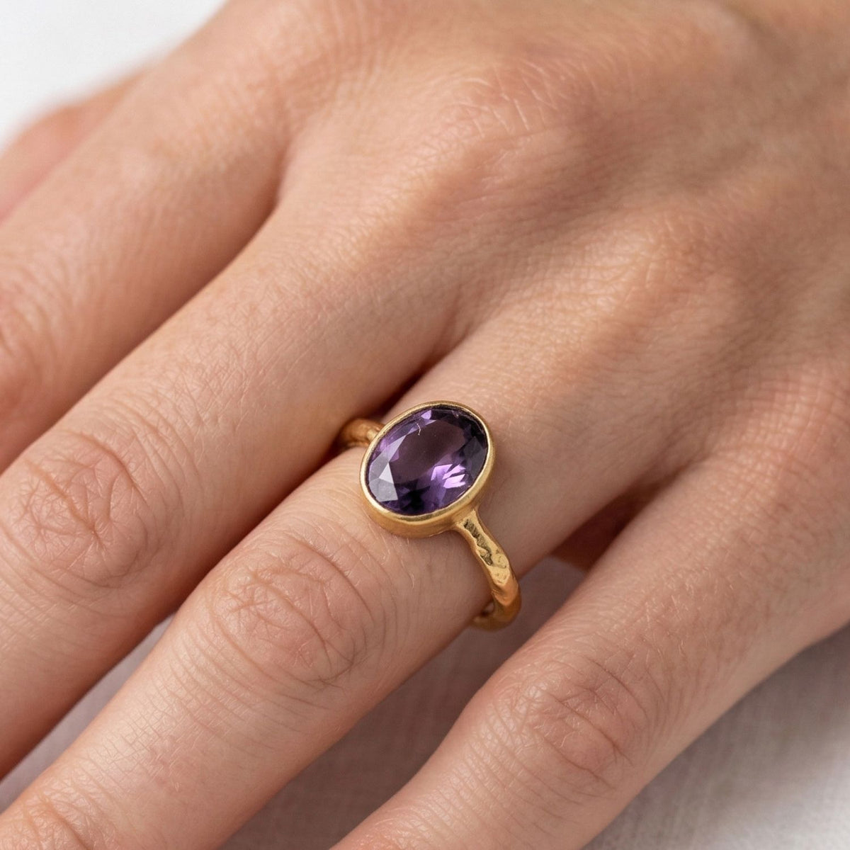 A hand with natural nails wears the JAREDJAMIN Jewelry Online Oval Amethyst Gemstone Ring, featuring a faceted purple amethyst in a smooth bezel on a textured band. The photo highlights the ring finger and the design’s elegant simplicity.