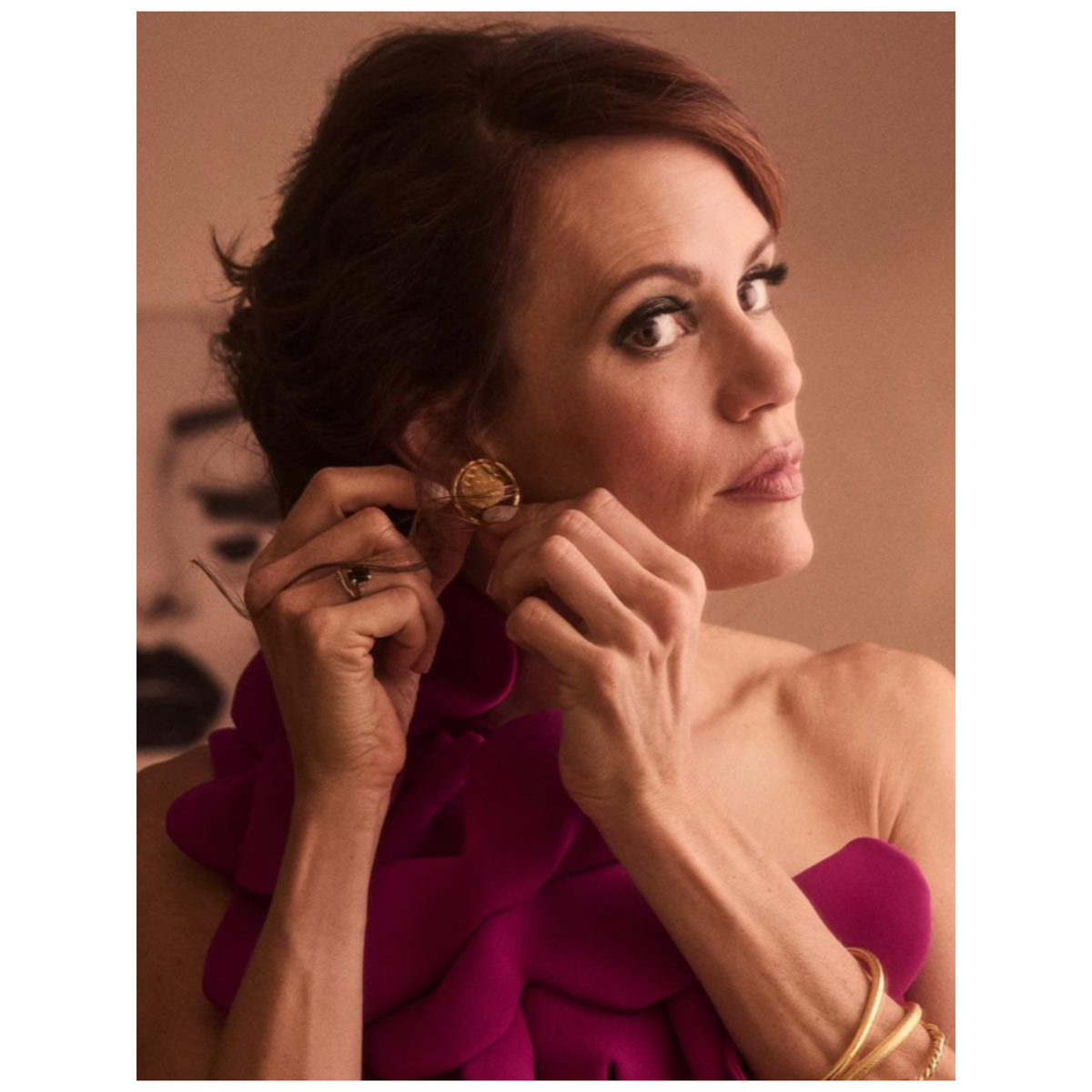 Un woman avec light skin et auburn hair styles her hair while wearing or Peacock Eye Post boucles d'oreilles de JAREDJAMIN Bijoux en Ligne, a magenta ruffled one-shoulder dress, et or joncs. Les background is softly blurred, avec abstract portrait details.