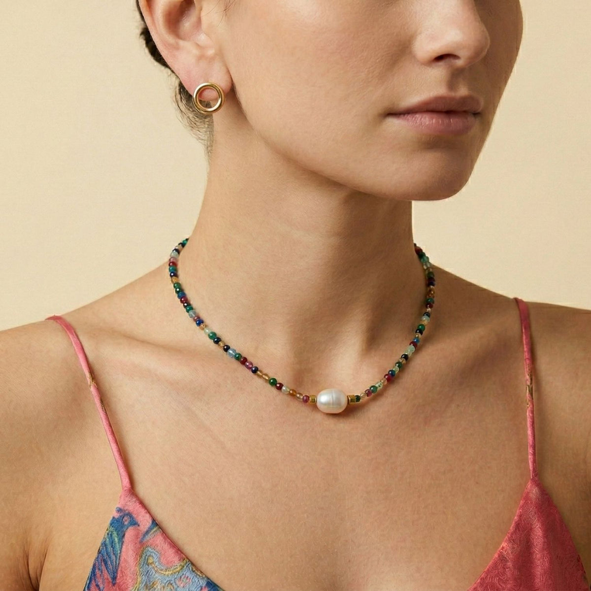 A woman wears the Rowan Multi Color Necklace from JAREDJAMIN Jewelry Online, featuring adjustable freshwater pearls, multi-colored faceted beads, and gold accents. She pairs it with gold hoops and a pink printed spaghetti strap top.