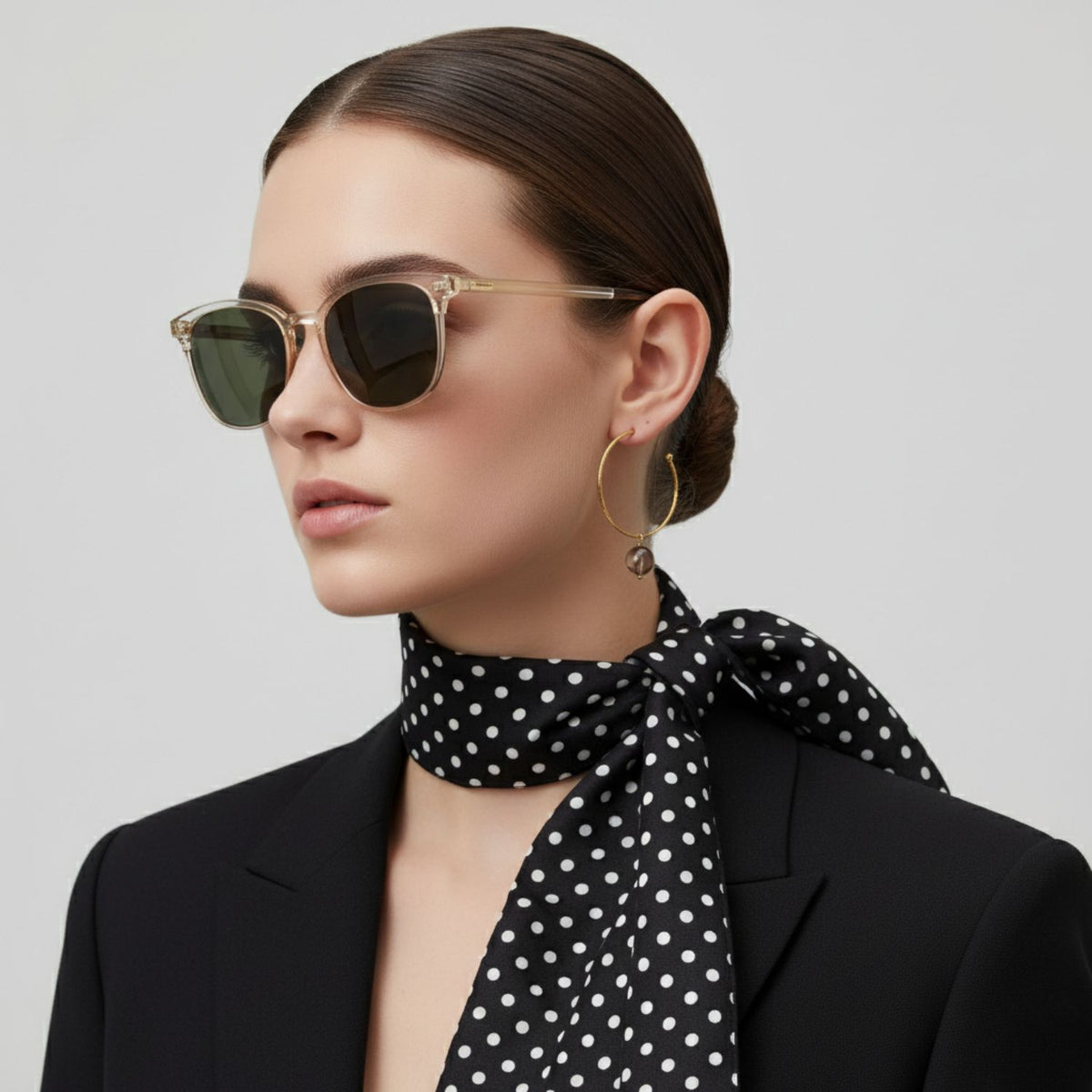 Un woman avec slicked-back hair wears round sunglasses, a black blazer, et a polka dot scarf. She showcases the 16MM Smooth Ball boucle d'oreille breloques par JAREDJAMIN Bijoux en Ligne against a plain light grey background.