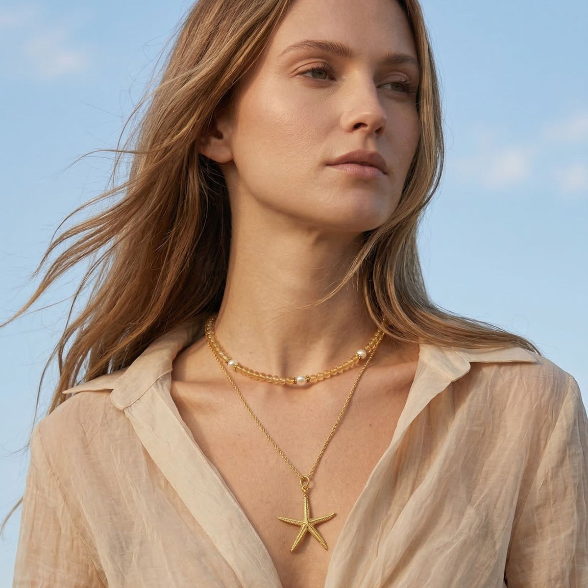 Los Sand Bar Sensation Citrine collar de JAREDJAMIN Joyería Online takes center stage, worn over a sheer beige blouse. Los model gazes calmly off-camera, framed by a clear, pale blue sky, con long light brown hair visible.
