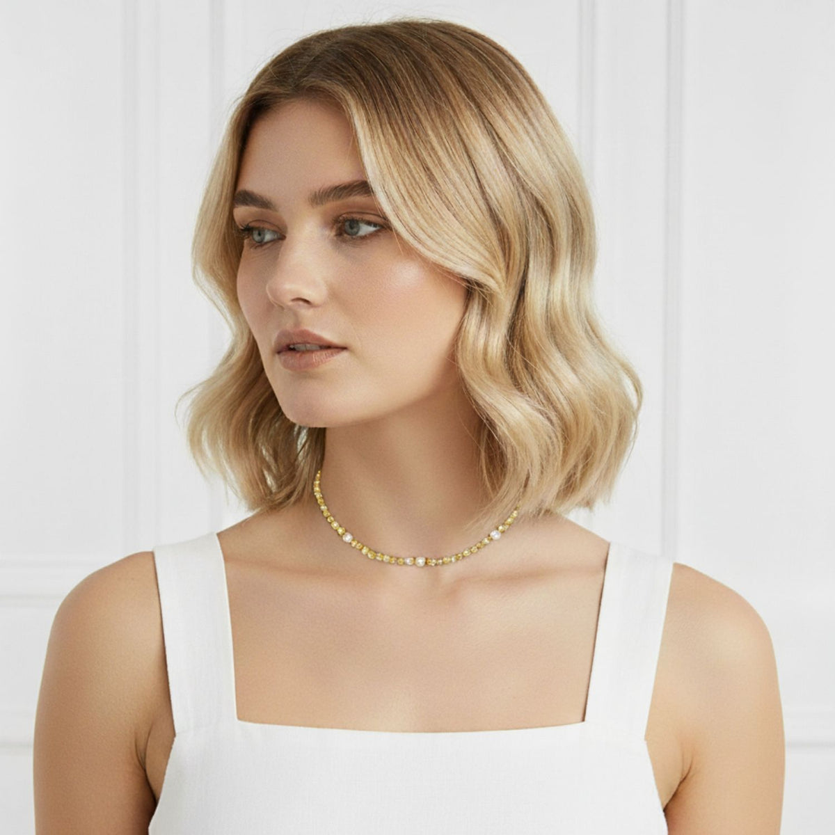 A woman with chin-length, wavy blonde hair and fair skin wears a square-neck white top and the Sand Bar Sensation Citrine Necklace from JAREDJAMIN Jewelry Online. She looks left calmly against a white, paneled background.