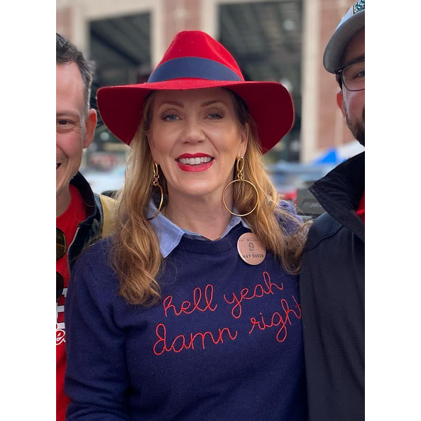Un smiling woman avec long blonde hair wears a red-brimmed hat, a navy “hell yeah damn right” sweater avec a button, et JAREDJAMIN Bijoux en Ligne's or Ophelia pendante créole Post boucles d'oreilles. She stands between two people; a building is blurred in the background.