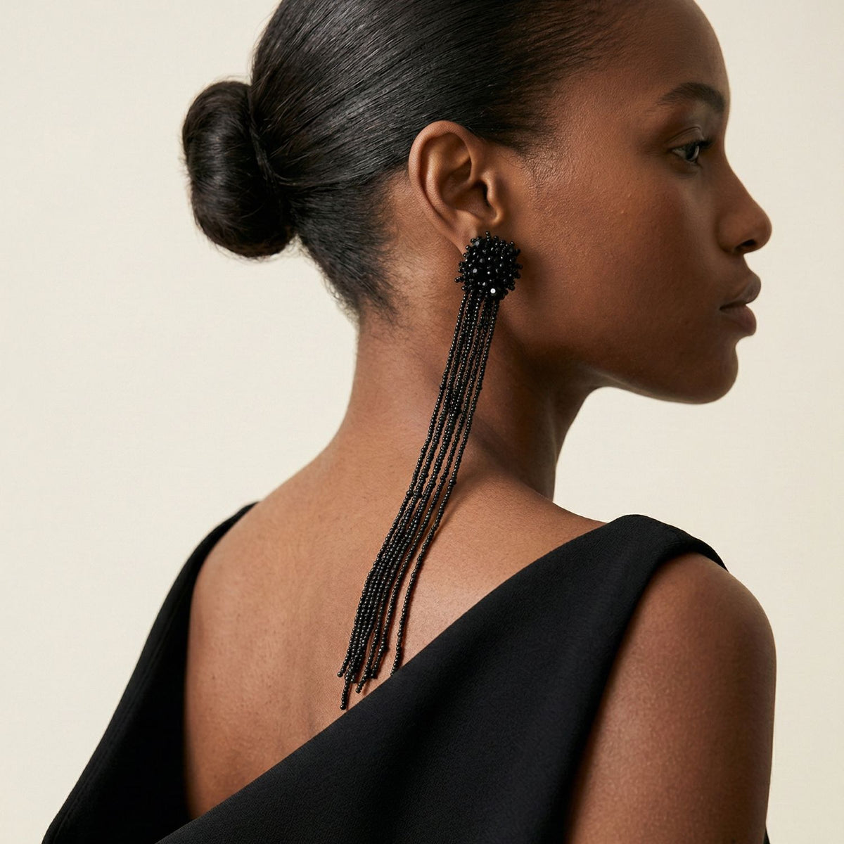 A woman with smooth dark skin and a neat low bun wears a sleeveless black dress with a deep back. She models JAREDJAMIN Jewelry Online’s Serafina Beaded Clip Earrings, their black glass beads dangling past her shoulder against a plain background.