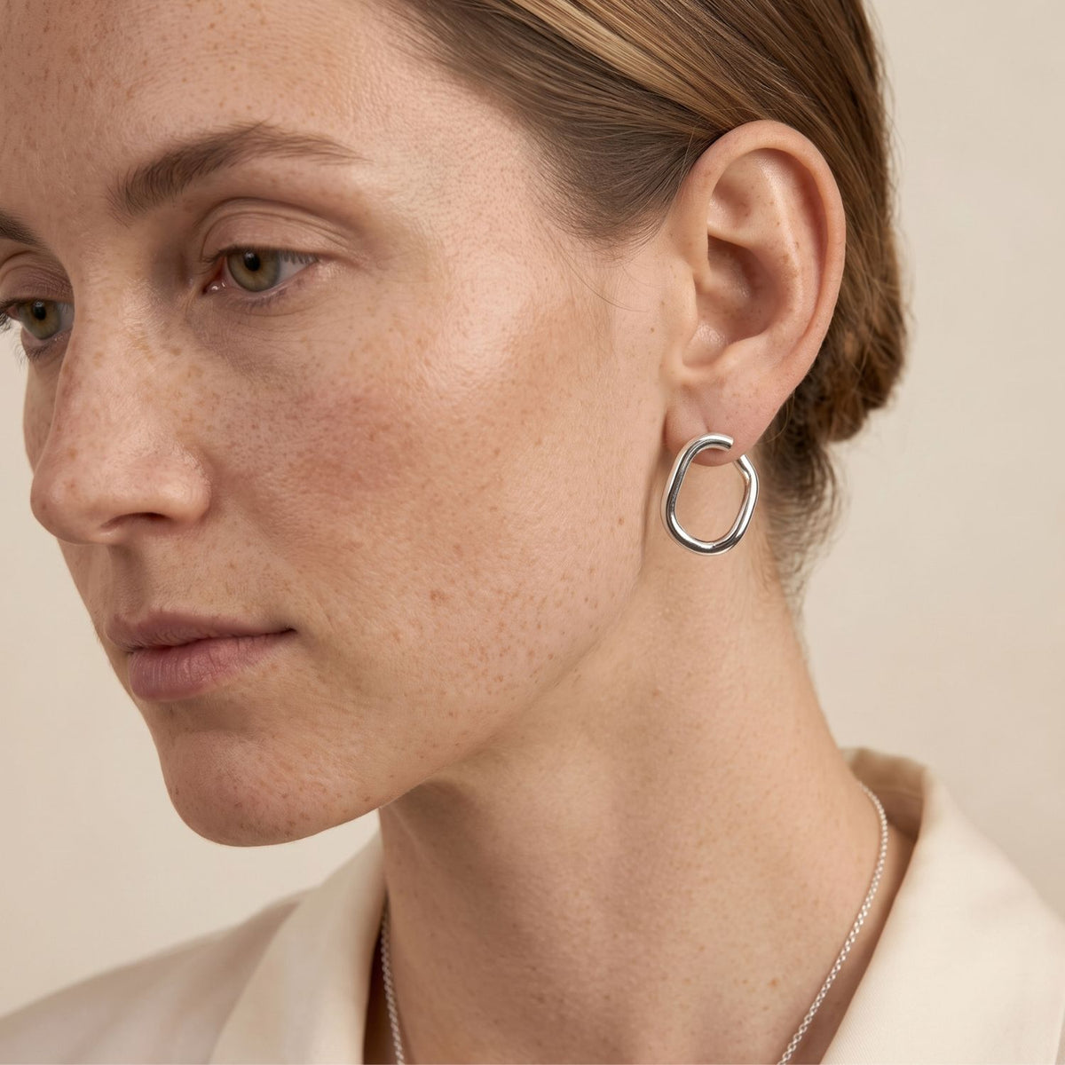 Close-up profile of a model's ear styling the 1-inch Silver Billie Stud Post Earrings, showcasing how the open silver oval link creates a striking, liquid-metal floating effect against the skin.