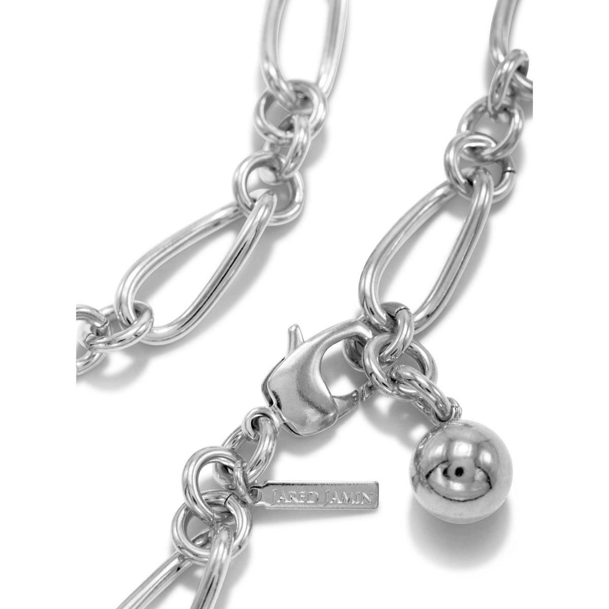 gros plan of the argent Skyler chaîne bracelet par JAREDJAMIN Bijoux en Ligne, avec brillant argent elongated oval links, a lobster clasp, a round metallic bead breloque, et a branded tag resting sur un fond blanc.