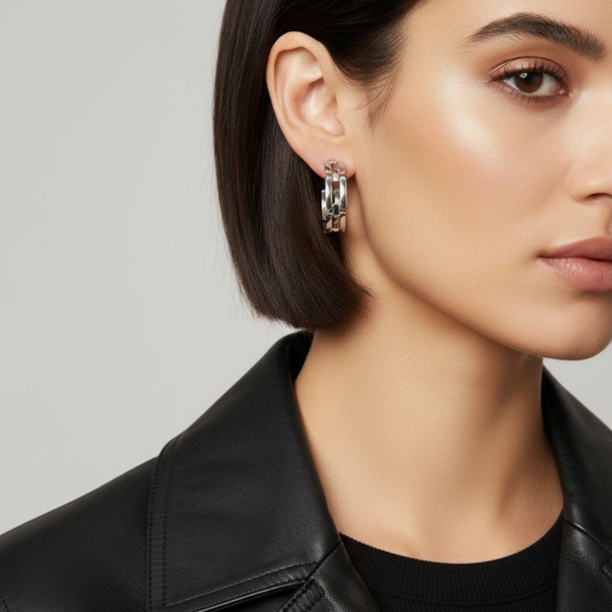 Un woman avec straight, chin-length dark hair wears a black leather jacket et JAREDJAMIN Bijoux en Ligne’s argent Bravura chaîne créole Post boucles d'oreilles, their platinum shine enhancing her glowing skin et natural makeup.