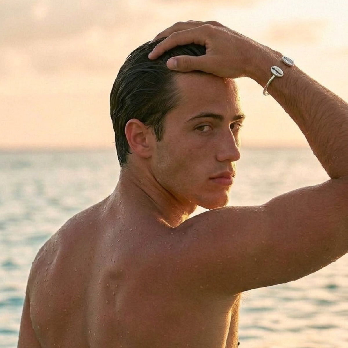 Editorial portrait of a male model in the ocean at sunset, running a hand through his hair while wearing the unisex Silver Cowrie Shell Cuff Bracelet on his wrist.