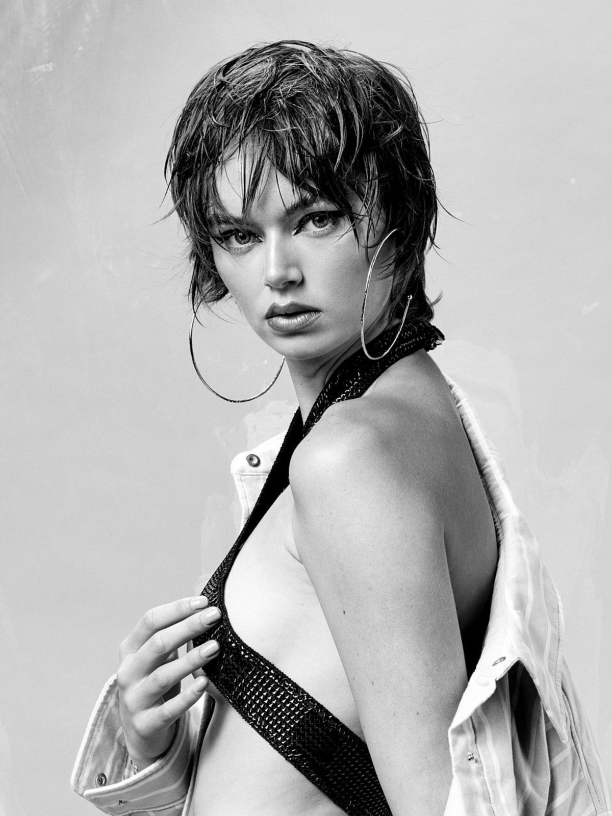 A black and white photo shows a person with short, wet hair wearing Silver Large Hooptastic Hoop Post Earrings by JAREDJAMIN Jewelry Online. With a confident expression, they gaze at the camera as a sequined strap crosses their bare shoulder and a jacket drapes over one arm against the plain light background.