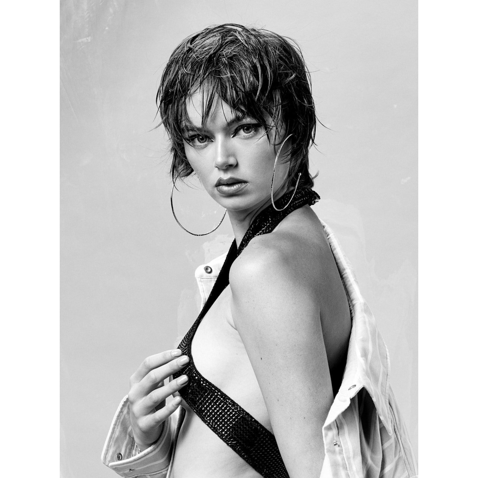 Black and white photo of a woman with short wet-look hair, wearing JAREDJAMIN Jewelry Online's Silver Large Hooptastic Hoop Post Earrings and a sparkly halter top, gazing seriously at the camera while holding her jacket off her shoulder.