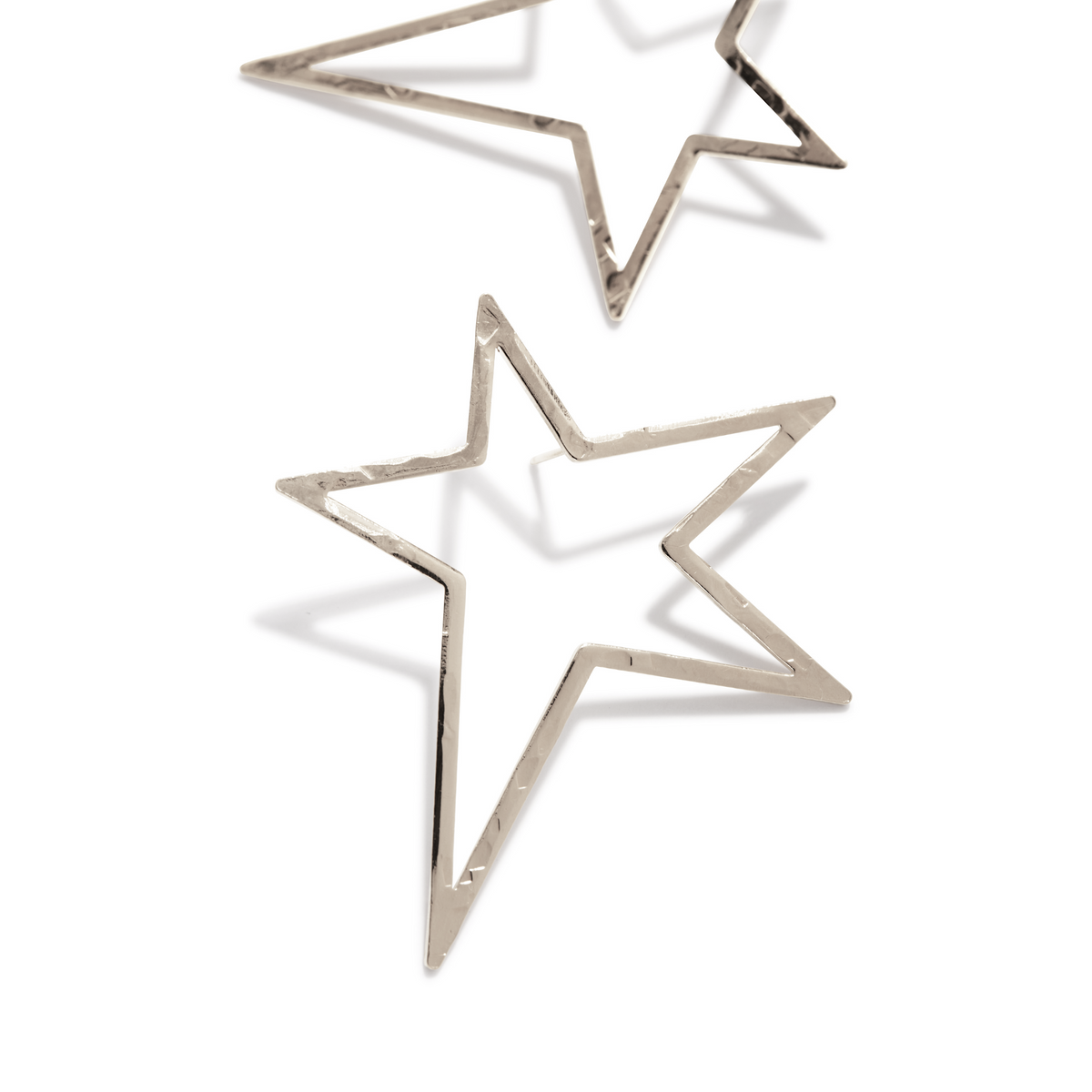Los plata Shooting Star Dust Post pendientes de JAREDJAMIN Joyería Online presentan large, plata-plated, hammered star outlines displayed diagonally sobre un fondo blanco, con soft shadows beneath them.