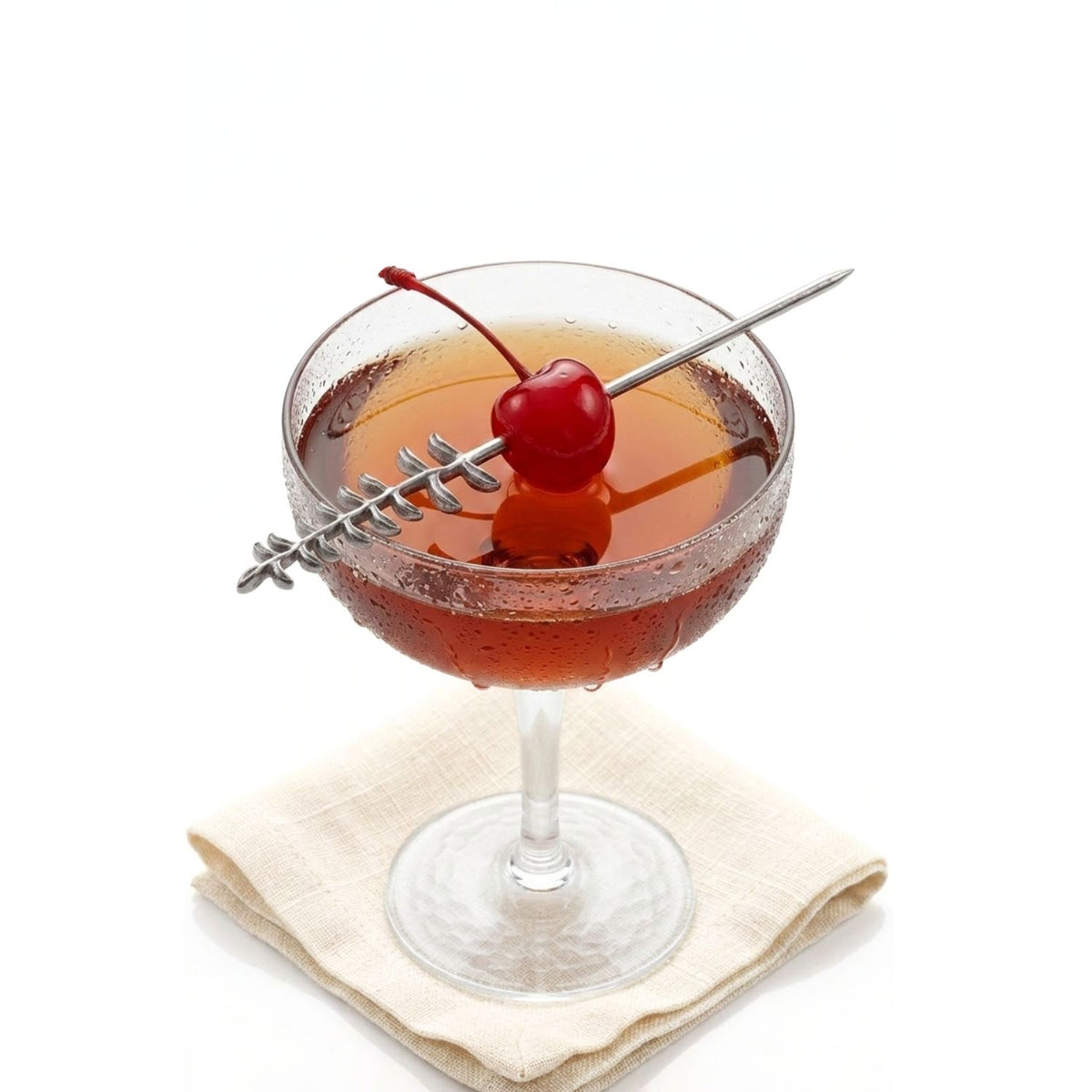 Luxury mixology styling shot of a clear crystal coupe glass holding a dark cocktail, with a 5 3/4-inch Silver Thyme Cocktail Skewer resting horizontally across the rim, piercing a bright cocktail cherry to demonstrate its botanical design and functional scale.