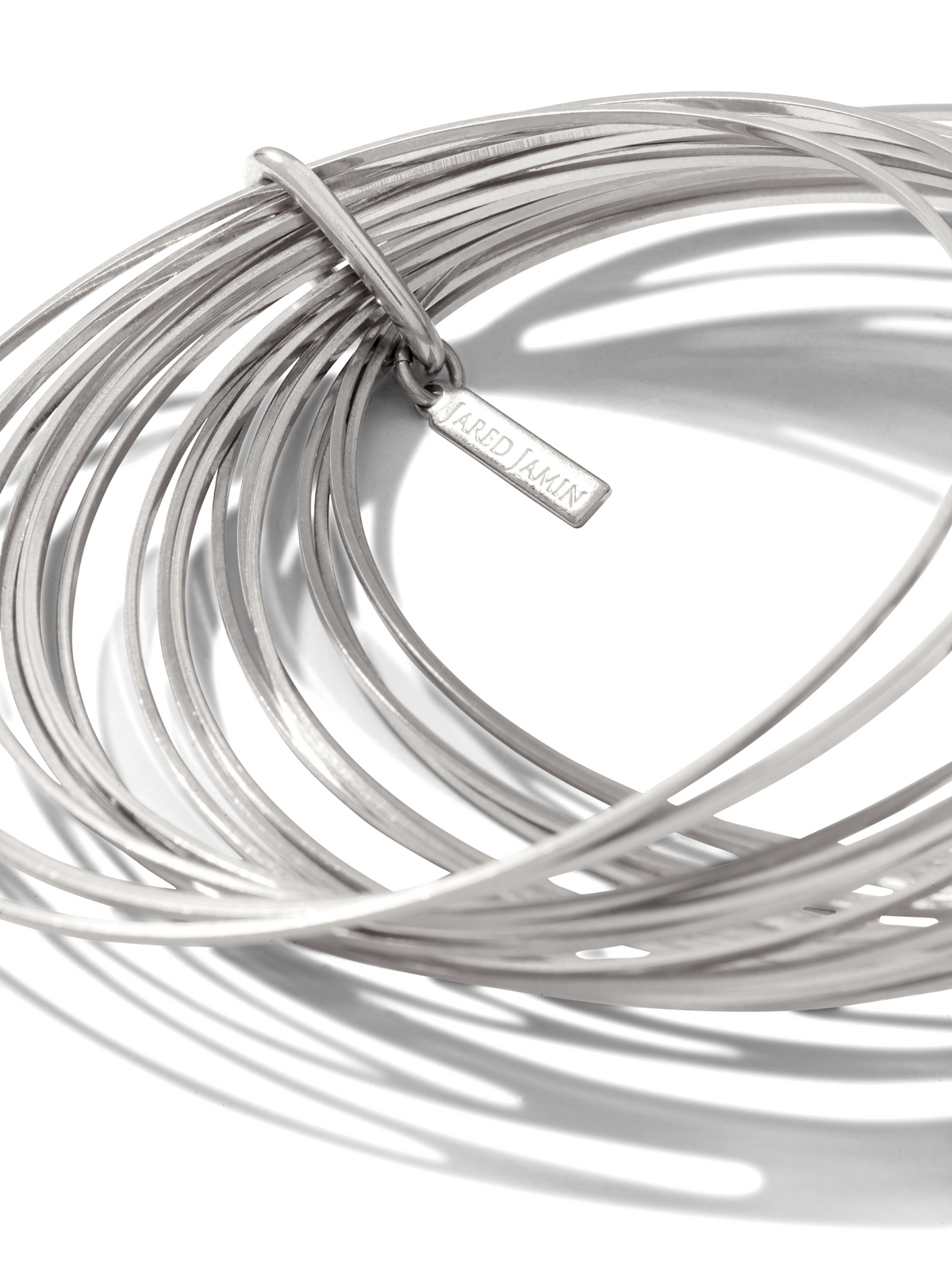 Un gros plan of JAREDJAMIN Bijoux en Ligne’s argent Twenty Something jonc Stack shows slim, argent-plated joncs stacked avec a branded rectangular tag, their metallic sheen et sleek design highlighted against a fond blanc.