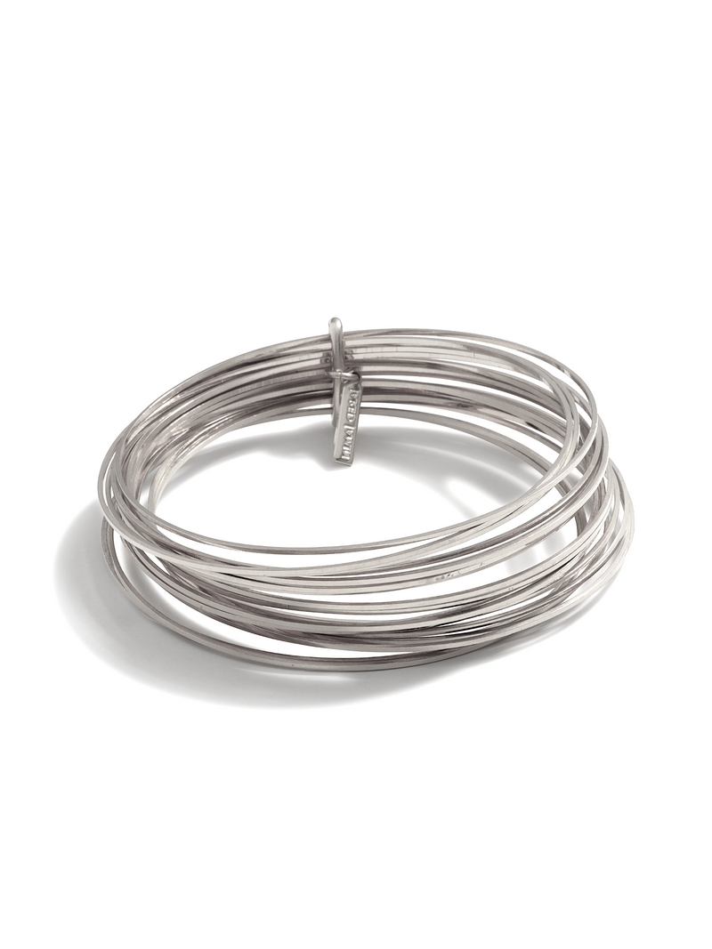 The Silver Twenty Something Bangle Stack from JAREDJAMIN Jewelry Online features seven slim, silver-plated bangles interlocked by a rectangular bar engraved with the brand name, all with a smooth, polished finish.