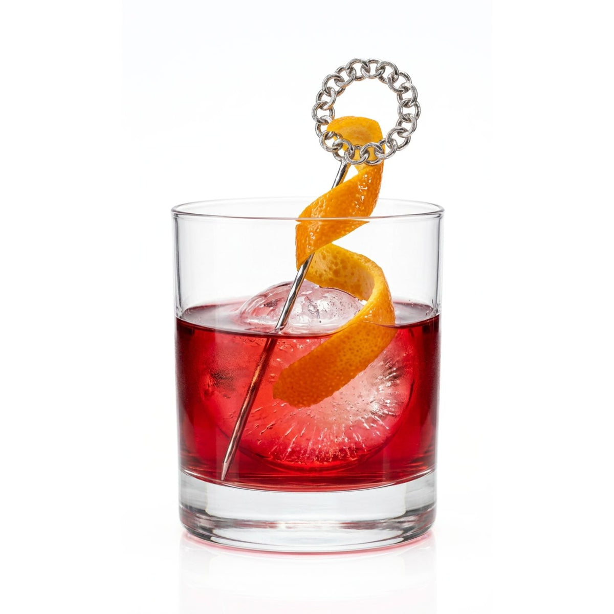 Luxury mixology styling shot of a clear crystal rocks glass holding a vibrant amber cocktail with a large ice sphere, showing a 4 7/8-inch Silver Wreath Cocktail Skewer standing vertically inside to anchor an elegant, long orange peel twist garnish.