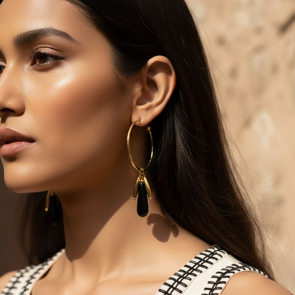 A woman with long dark hair wears JAREDJAMIN Jewelry Online’s Small Hooptastic Wisteria Charm Gold Hoop Earrings, featuring removable teardrop charms, paired with a sleeveless patterned top. The close crop highlights her left profile in soft sunlight.