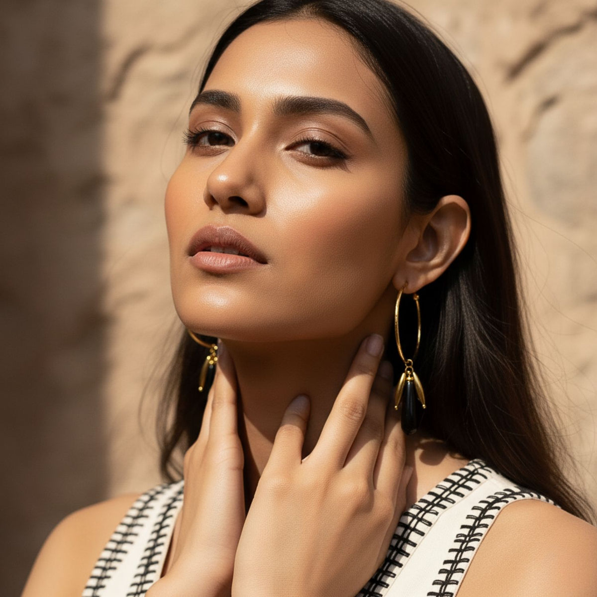 A woman with long, dark hair poses by a sunlit stone wall, wearing JAREDJAMIN Jewelry Online's Small Hooptastic Wisteria Charm Gold Hoop Earrings and a sleeveless white top with black stitching. Her glowing skin shines as she gazes confidently at the camera.