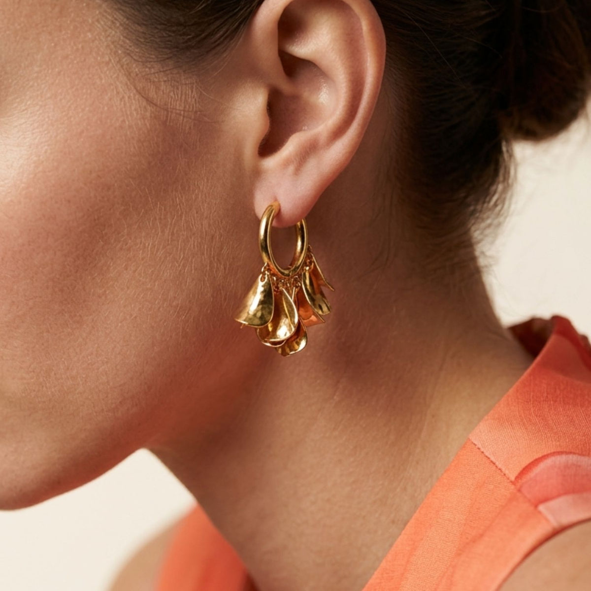 Side profile portrait view of a model wearing the 1.25-inch Small Petali Cascading Flower Hoop Earrings, showing their volumetric botanical shape and reflective gold finish styled with a peach coral top.
