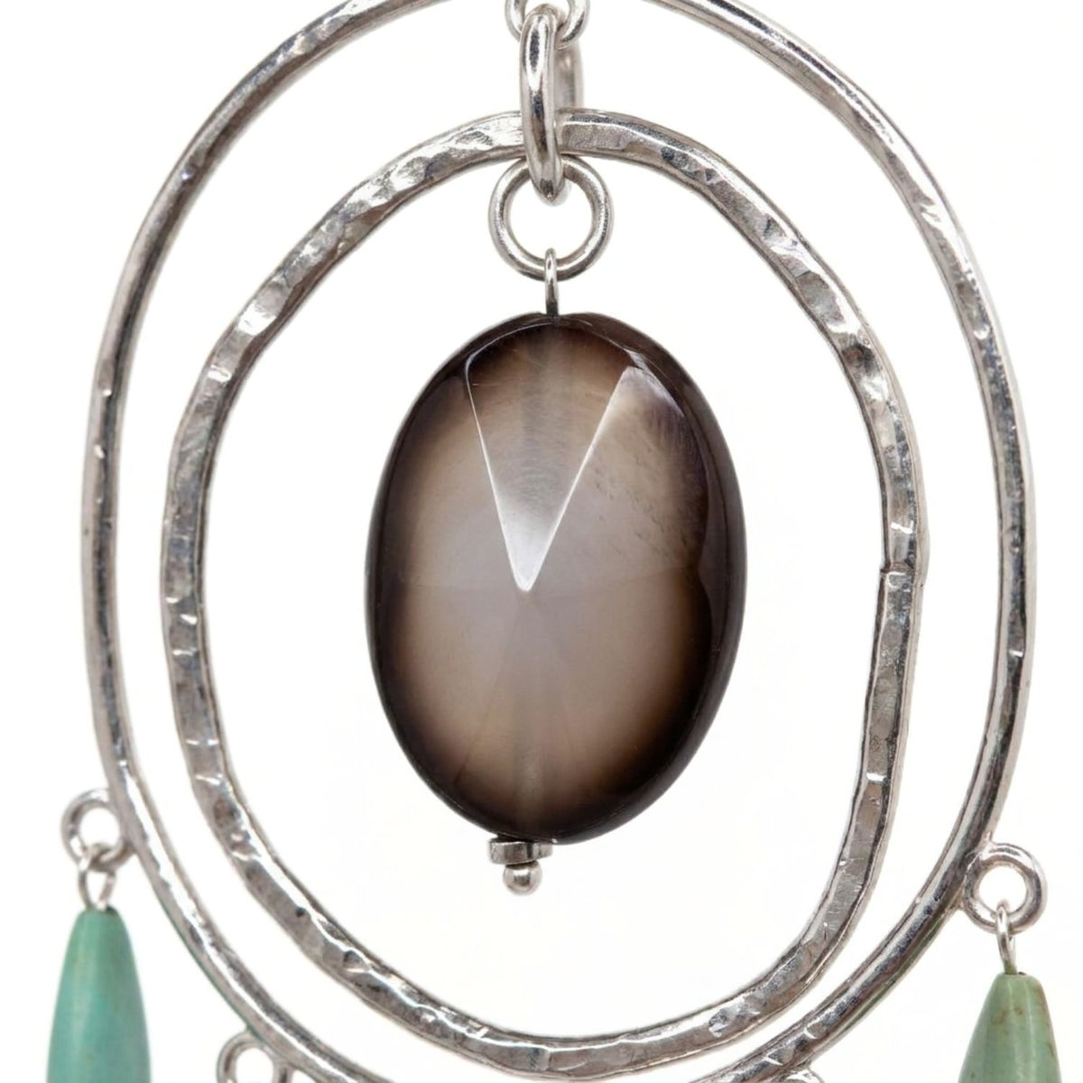 Close-up studio detail shot focusing on the focal faceted oval agate bead with an opaque center and dark brown rim, suspended inside hand-hammered silver-plated brass concentric wire rings with lower turquoise howlite charms.