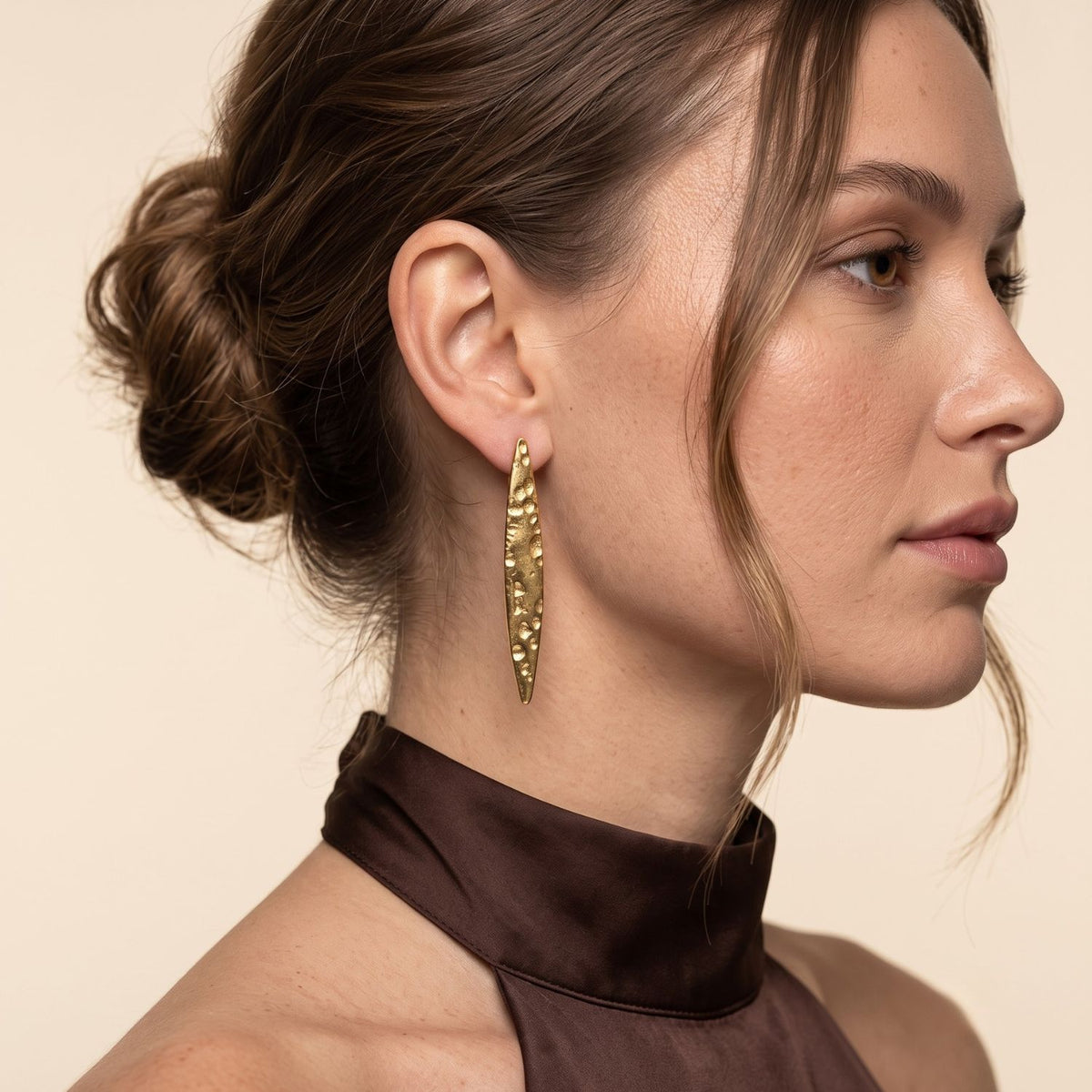 Side profile portrait of a model wearing the 2-inch 18k gold-plated Soren hammered marquise post earring, showcasing its light-diffusing dimpled surface and vertical drop.