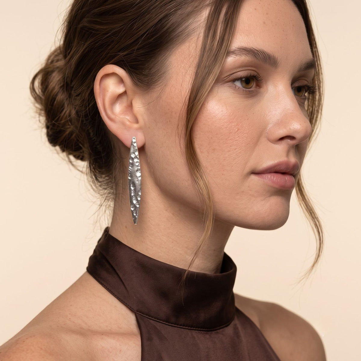 Side profile portrait of a model wearing the 2-inch silver-plated Soren hammered marquise post earring paired with a brown satin halter top, demonstrating its flush fit along the lobe.