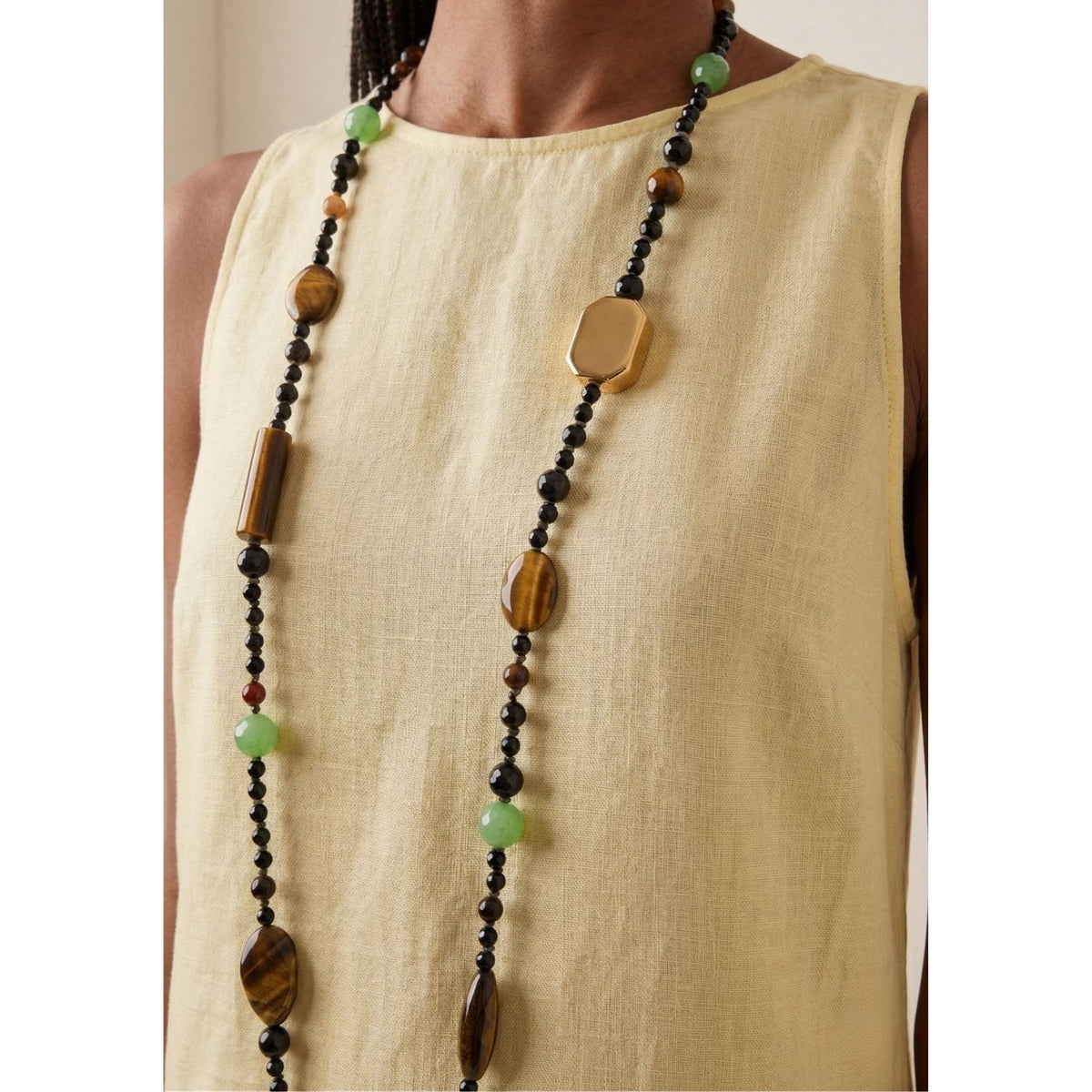 A woman in a sleeveless light yellow linen top wears the Sorrento Gemstone Knotted Necklace—hand-assembled in our West Hollywood studio from earthy, gold, black, and green beads. Crafted for small-batch impact with eco-conscious intent. Style Note: punctuates day-to-night silhouettes.