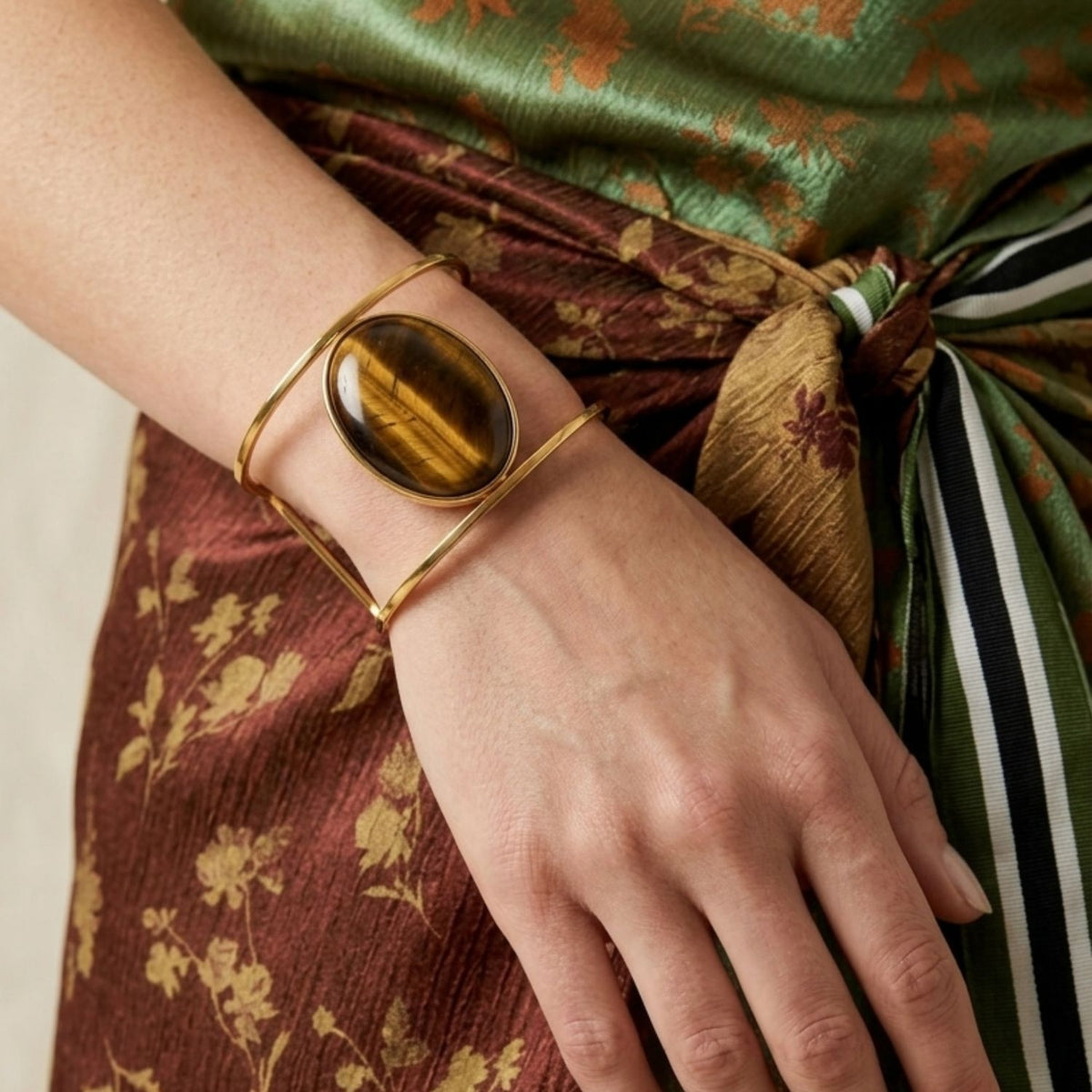 An 18K gold-plated Stella Cuff bracelet with a bold oval Tiger’s Eye stone adorns the wrist, drawing focus over the resting hand on a floral brown skirt tied with a green, white, and black striped sash and a green-gold top.
