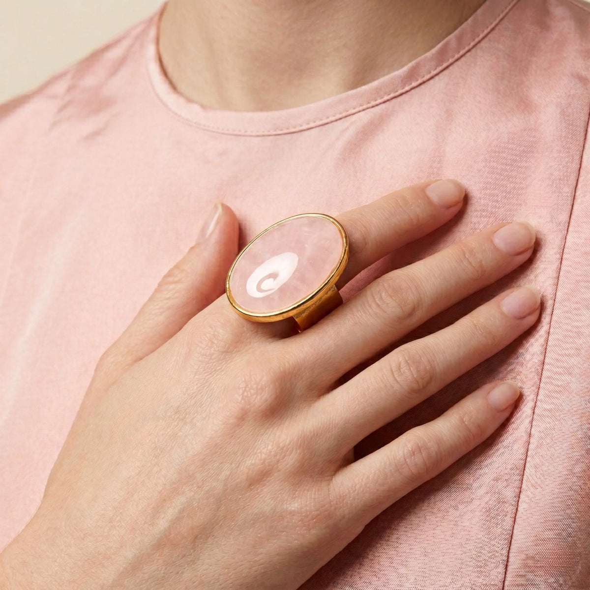 A person in a pale pink satin top rests their hand on their chest, punctuating the moment with the Stella Statement Ring in Rose Quartz—an 18k gold-plated, hand-assembled centerpiece sculpted in our West Hollywood studio. Small-batch, eco-conscious. Style Note: shifts from polished daywear to after-dark refinement. Nails are neatly manicured and natural.
