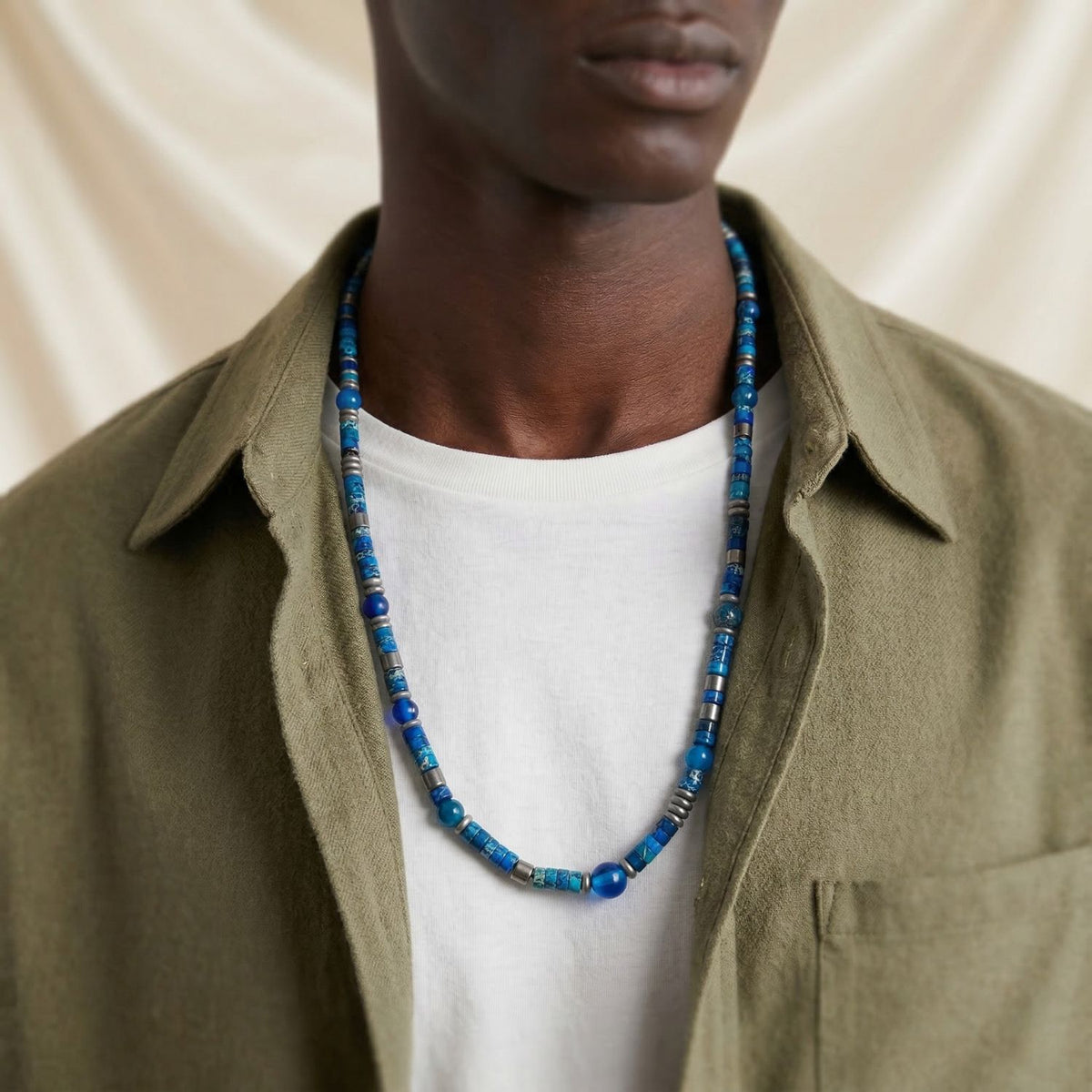 JAREDJAMIN bijoux Surf Side Jasper Beaded collier avec faceted agate beads et argent-plated hematite accents is shown against a beige fabric backdrop. Les collier is modeled over an olive green shirt layered sur un white tee.