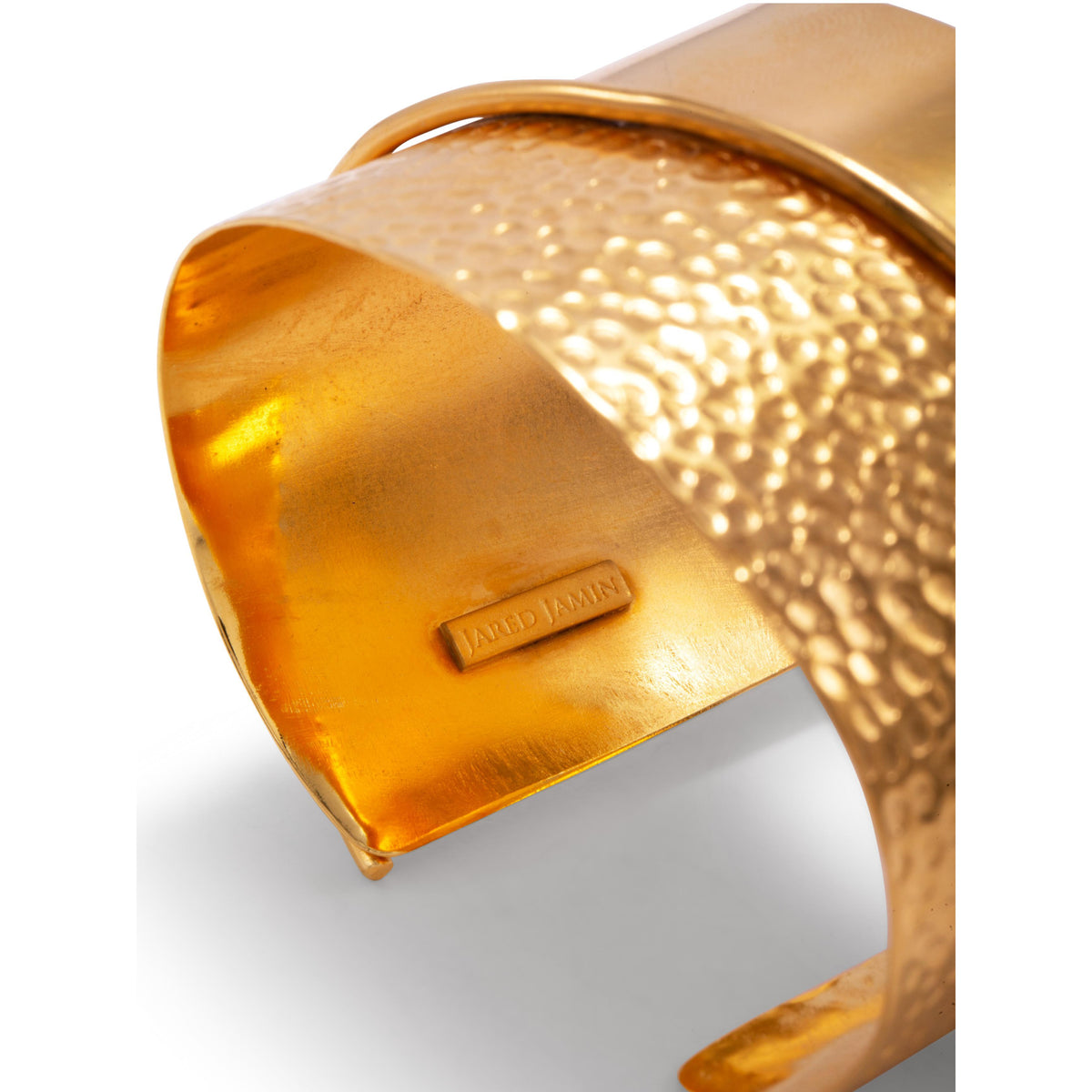 Un primer plano of the Tribella oro puño pulsera de JAREDJAMIN Joyería Online shows its wide, hammered 18K oro-finished band y a small inner tag engraved con “Jared Jamin,” all set against a plain fondo blanco.