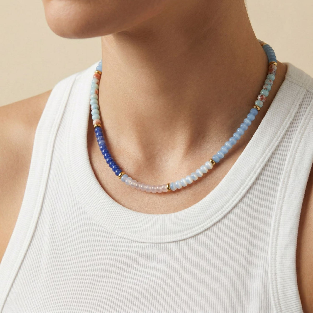 A woman models the Taylor Beaded Necklace Blue, meticulously hand-assembled in our West Hollywood studio. Light blue, white, and orange beads punctuate her neckline—crafted in small, eco-conscious batches. Style Note: layers seamlessly from tanks to tailored blazers.