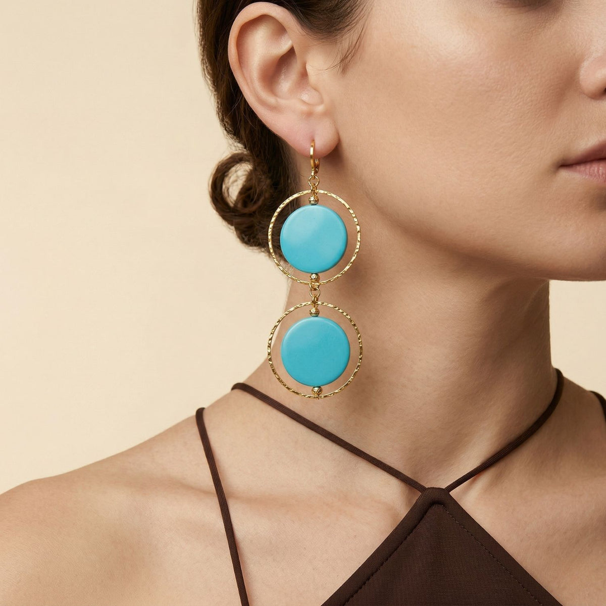 A woman in a brown halter-neck top wears JAREDJAMIN’s Twiggy Turquoise Dangle Earrings—turquoise-blue discs orbiting textured gold hoops. Hand-assembled in our West Hollywood studio with small-batch, eco-conscious intent. Style Note: transitions from sharp tailoring to silk after dark. Her hair is pulled back against a beige backdrop.