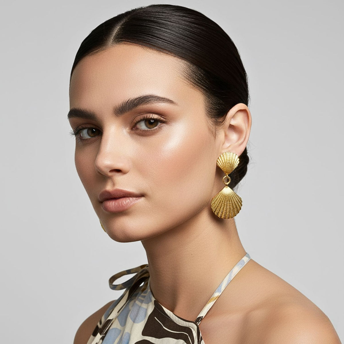 A woman with smooth, dark hair in a low bun wears the Gold Venus Clip On Earrings by JAREDJAMIN Jewelry Online. She has glowing skin and defined brows, and her halter top features a geometric pattern in brown, blue, and cream.