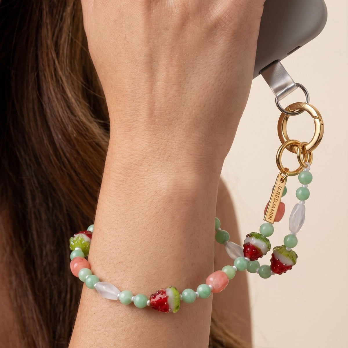 Close-up lifestyle shot of a model's wrist holding a smartphone, with the 16-inch Watermelon Crush strand wrapped multiple times as a structured gemstone bracelet, highlighting its multi-wear utility and gold hardware attachment.