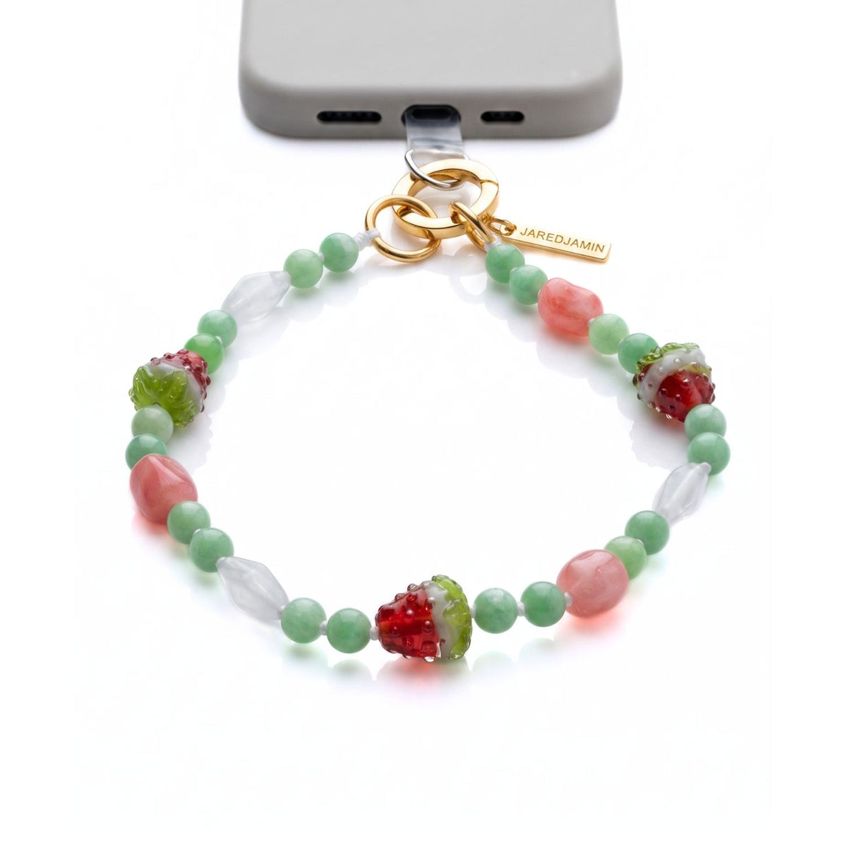 High-end studio shot of the JAREDJAMIN Watermelon Crush Strap Wristlet attached to a gray smartphone case via a clear anchor tab, showing the hand-knotted watermelon quartz, clear rock crystal, and glass watermelon slice charms connected to an 18k gold-plated circular clicker push clasp.