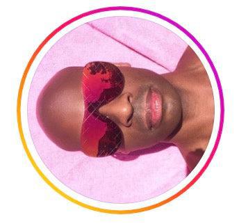 A person with a shaved head is lying down on a pink surface, wearing large, heart-shaped sunglasses with red lenses that reflect the sky. Their lips are slightly parted, and the image is framed with a colorful gradient border.