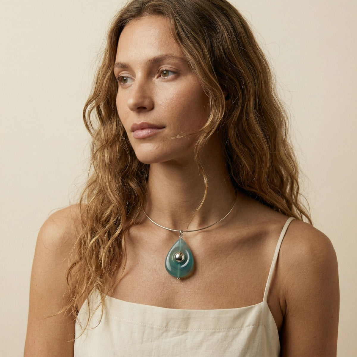 A woman with long, wavy light brown hair in a cream spaghetti-strap top wears the hand-assembled Zenith Pendant—crafted in our West Hollywood studio. Made small-batch with eco-conscious methods. Style Note: punctuates both soft knits and sharp tailoring.