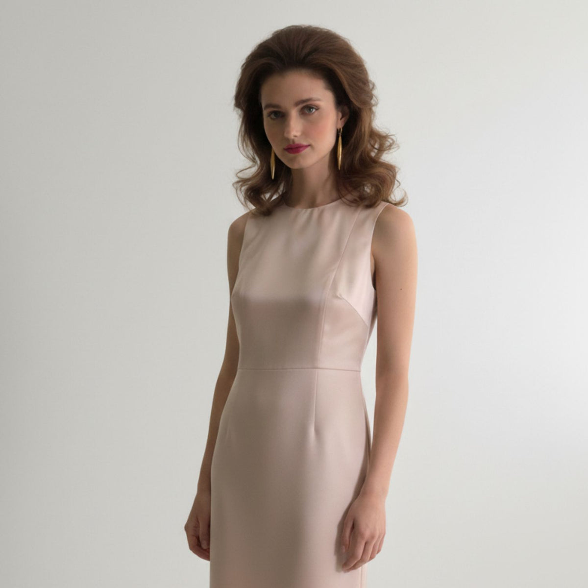 A woman with wavy brown hair wears a sleeveless, light pink fitted dress and stands against a plain background with a calm expression, accessorized with JAREDJAMIN Jewelry Online&#39;s Zeppelin Cora 1 Drop Gold Earrings.