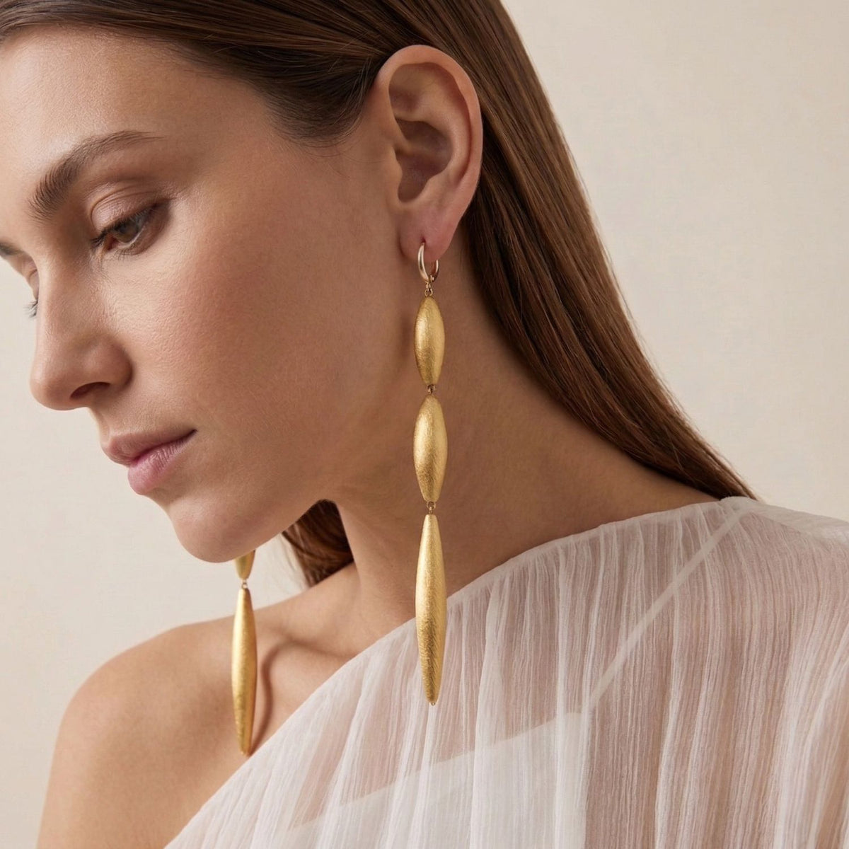 A woman with straight brown hair and soft, natural makeup wears a sheer white, one-shoulder top. Zeppelin Cora 3 Drop Gold Leverback Earrings—hand-assembled in our West Hollywood studio—punctuate the look: eco-conscious, small-batch, endlessly versatile from work to evening.