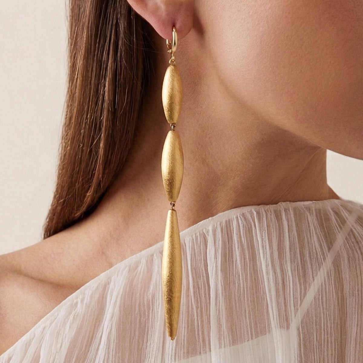Zeppelin Cora 3 Drop Gold Leverback Earrings by JAREDJAMIN showcase matte-finish, oval and elongated forms. Crafted in small batches West Hollywood, these sustainable luxury earrings feature brushed textures—JAREDJAMIN Essentials. Seen with a sheer white top.